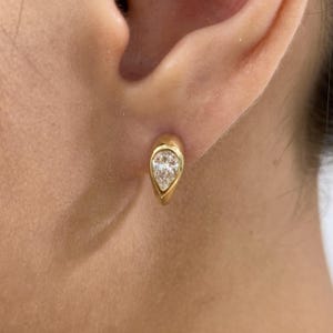 May include: A gold earring with a pear-shaped diamond set in a bezel setting. The earring is a stud style, and the diamond is clear and sparkling. The earring is worn on an ear.