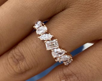 May include: A rose gold band ring featuring a series of clear, faceted stones in various cuts, including round, marquise, and emerald shapes, arranged in a row across the top of the band, creating a multi-faceted, sparkling appearance.