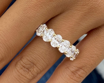 May include: A gold band ring featuring a row of five clear oval-cut stones set in prongs. The stones are arranged horizontally along the finger, showcasing a brilliant faceted surface that reflects light across the entire setting of the jewelry piece.