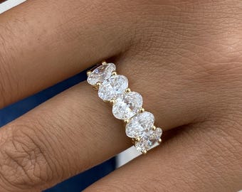May include: A gold band ring featuring five oval-cut, clear gemstones. The gemstones are set closely together, creating a continuous row across the top of the ring. The ring is shown on a person's finger.