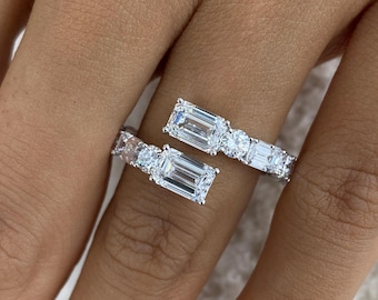 May include: A bypass ring crafted from silver-toned metal featuring two emerald-cut clear stones at the open ends, each flanked by a round brilliant-cut stone and additional smaller rectangular stones along the band's shank.