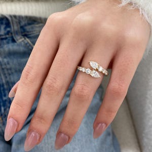 以下が含まれることがあります： ゴールドトーンのリング。バイパスデザインで、丸型とマーキス型の透明な宝石が並んでいます。指に巻き付くデザインで、モダンでエレガントな印象です。宝石はゴールドのバンドの上で輝いています。