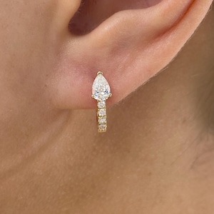 May include: A gold hoop earring with a pear-shaped diamond and small, round diamonds. The earring is worn on an ear, showcasing the jewelry's design and sparkle. The diamonds are clear and reflect light.