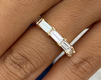 May include: A gold band ring featuring a row of four clear, rectangular baguette-cut stones set in a prong setting. The metal band has a polished finish, and the stones are arranged horizontally along the top section of the ring.