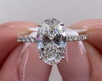 May include: A silver-toned ring featuring a large, oval-cut center stone held by eight prongs. The thin band is encrusted with a series of small, round, clear stones along the sides, creating a sparkling, reflective appearance on the metal surface.