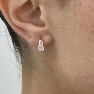 May include: A gold-colored earring with a pear-shaped, clear gemstone. The earring is a small hoop style, with the gemstone set within the hoop. The earring is worn on the ear.