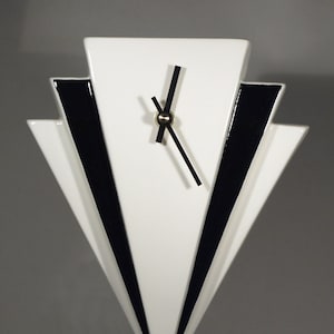 Könnte beinhalten: Eine weiße Keramikuhr mit schwarzen Akzenten in Form eines Art-Deco-Fächers. Die Uhr hat schwarze Zeiger und ein goldenes Zentrum.