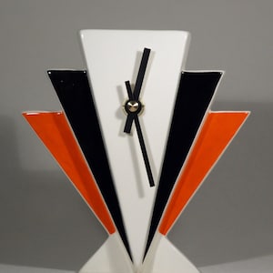 Könnte beinhalten: Eine geometrische Uhr im Art-Deco-Stil mit weißem Zifferblatt und schwarzen Zeigern. Die Uhr hat ein weißes, schwarzes und orangefarbenes Design. Die Zeiger der Uhr sind schwarz mit einem goldfarbenen Zentrum. Die Uhr ist ein dekorativer Einrichtungsgegenstand.