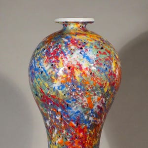 Puede incluir: Un jarrón decorativo de cerámica con cuerpo bulboso y base acampanada. El jarrón está cubierto con un colorido patrón de salpicaduras de pintura roja, azul, naranja, amarilla y negra. El borde es blanco.