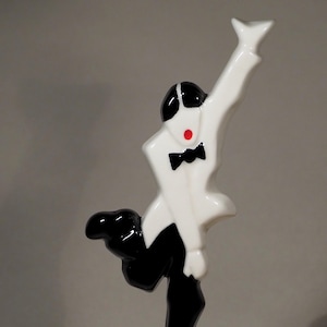 May include: A glossy ceramic figurine of a figure in a white jacket and black trousers, wearing a black bow tie and sporting a red dot on the face. The figure is posed with one arm raised and one leg bent, standing on a red base atop a white plinth.