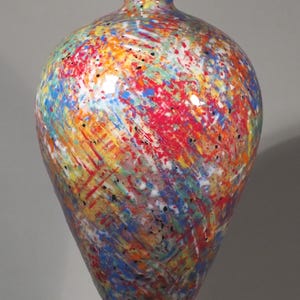Puede incluir: Un jarrón de vidrio con cuerpo bulboso y cuello estrecho que presenta un vibrante patrón de salpicaduras multicolor. La superficie muestra rayas de pintura roja, azul, amarilla, naranja y verde sobre una base transparente, con un borde blanco liso.