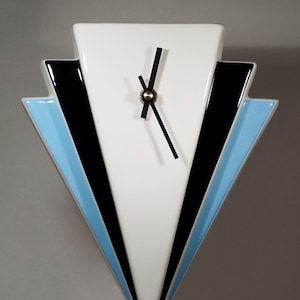 Könnte beinhalten: Eine geometrische Wanduhr mit weißem Zifferblatt, schwarzen Zeigern und einem goldfarbenen Zentrum. Die Uhr hat eine dreieckige Form mit schwarzen und hellblauen Akzenten. Das Design der Uhr erinnert an den Art-Deco-Stil.