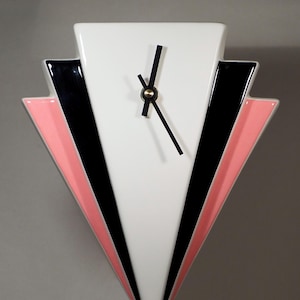 Könnte beinhalten: Eine geometrische Wanduhr mit weißem Zifferblatt und schwarzen Zeigern. Die Uhr hat eine dreieckige Form mit schwarzen und rosa Akzenten. Das Design der Uhr erinnert an den Art-Deco-Stil.