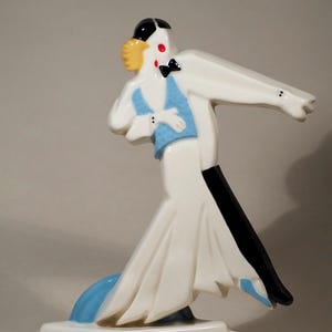 May include: A ceramic figurine of a dancing couple in Art Deco style. The man is in a black tuxedo with a bow tie, and the woman is in a white dress with a blue vest. The base is white, and the figurine has a glossy finish.