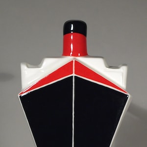 Puede incluir: Una escultura de cerámica blanca con forma de barco con un diseño geométrico rojo y negro. La escultura tiene un fondo puntiagudo y una parte superior plana con una pequeña chimenea.