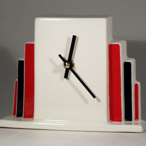 Può includere: Orologio in ceramica bianca con lancette nere e accenti geometrici rossi e neri. L'orologio ha un design semplice con un quadrante rettangolare e una base.