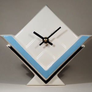 Pode incluir: Um relógio de cerâmica branca com um design geométrico. O mostrador do relógio tem formato de diamante com ponteiros pretos e um centro dourado. Possui uma faixa azul clara e detalhes em preto. O relógio mede cerca de 20 cm de altura.