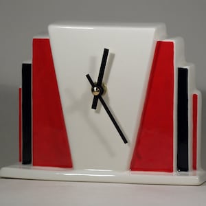May include: A white ceramic clock with black hands and red and black geometric designs. The clock has a simple, modern design.