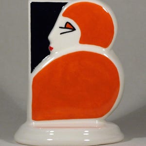 Könnte beinhalten: Keramikfigur mit stilisiertem Frauenprofil in Orange und Weiß. Die Figur hat einen schwarzen rechteckigen Hintergrund und eine rote Lippe. Der Sockel ist weiß und oval geformt. Die Figur ist ein dekoratives Kunstwerk.
