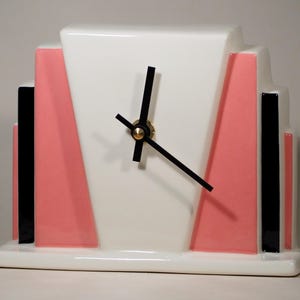 Könnte beinhalten: Eine keramische Art-Deco-Uhr mit weißem Zifferblatt und geometrischem Design. Die Uhr hat schwarze Zeiger, eine goldfarbene Mitte und rosa und schwarze Akzente. Die Uhr ist rechteckig und steht auf einem weißen Sockel.