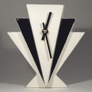 Pode incluir: Relógio de cerâmica em estilo Art Déco, creme e preto. O relógio apresenta um design geométrico com detalhes em preto e um mostrador dourado. Os ponteiros são pretos e o relógio tem formato triangular.