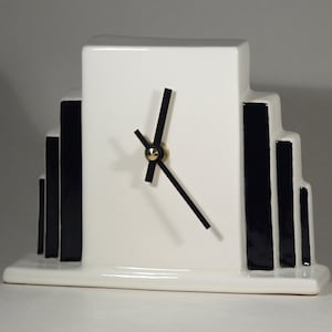 Könnte beinhalten: Weiße Keramikuhr mit schwarzen Zeigern und schwarzem geometrischem Design. Die Uhr hat einen weißen Sockel und ein rechteckiges Zifferblatt.