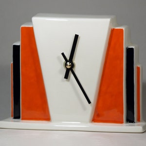 May include: A white ceramic clock with black hands and orange and black geometric accents. The clock has a modern art deco design.