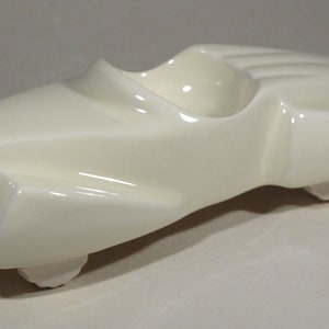 May include: A cream-colored, ceramic ashtray shaped like a vintage race car. The ashtray has a sleek, aerodynamic design with a rounded front, a cockpit-like depression, and a rear fin. It rests on small, rounded feet.