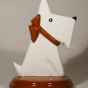 May include: A white ceramic Scottie dog figurine with a brown bow and base. The dog has a black nose and eye, and the bow is a contrasting color. The figurine is a decorative item.