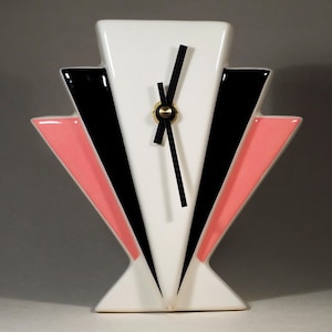 May include: Art Deco ceramic clock with a white face and black hands. The clock features a geometric design with black and pink accents. The clock has a gold-coloured centre and a rectangular base.