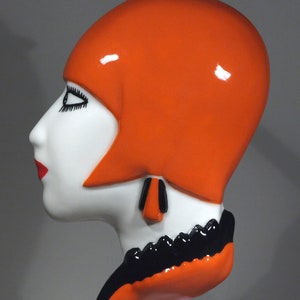 Op de afbeelding: Een keramische sculptuur van een vrouwenhoofd met een feloranje hoed met een zwarte en witte verenversiering. Het beeld is geschilderd in wit, zwart en oranje.