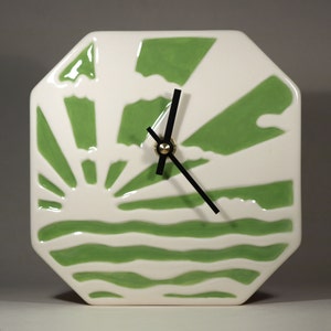 Puede incluir: Un reloj de cerámica blanca con un diseño de sol verde y blanco. El reloj tiene agujas negras y un centro dorado.
