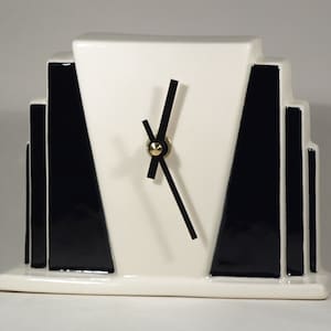 Könnte beinhalten: Eine weiße Keramikuhr mit schwarzen geometrischen Mustern und schwarzen Zeigern. Die Uhr hat ein goldfarbenes Zentrum.