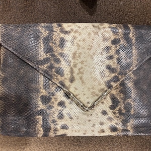 Puede incluir: Bolso de mano rectangular con estampado de piel de serpiente en tonos marrón, beige y negro. El cierre de solapa se asegura con un broche de metal plateado. El bolso está hecho de un material texturizado.