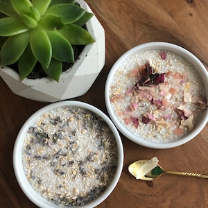 May include: Two white bowls filled with bath salts, one with dried lavender and oats, the other with rose petals and pink Himalayan salt. A gold spoon with a clear crystal sits on a wooden surface.