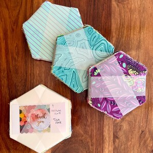 May include: Several hexagon-shaped fabric pieces with diverse patterns and colours, including stripes, swirls, and floral designs. One piece has a label with the words "thank you" and "medium 33 x 33 cm". The items are arranged on a wooden surface.