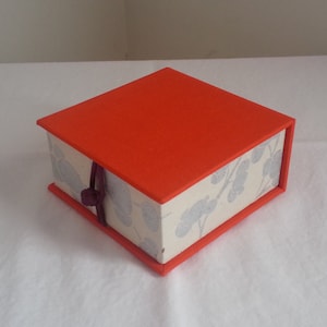Puede incluir: Una pequeña caja cuadrada con una tapa de color naranja brillante y una base blanca con un patrón floral. La caja tiene una cinta morada atada alrededor.