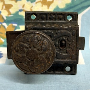 May include: A dark brown, cast iron rim lock featuring a circular knob with an embossed floral pattern. The rectangular base plate displays intricate scrollwork and a central keyhole, with mounting holes visible at the top and bottom edges.