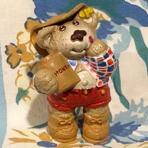 May include: A figurine of a bear wearing a hat, red shorts, and boots, holding a bag labeled "HONEY." The bear has a plaid shirt and a brown nose. The figurine is on a floral patterned background.