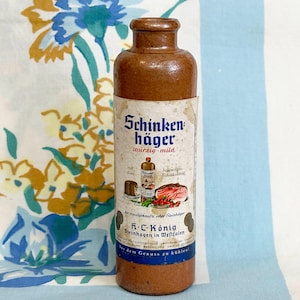May include: A brown ceramic bottle with a label featuring the text "Schinken häger würzig-mild". The label also includes illustrations of food items. The bottle is cylindrical with a narrow neck and is set against a floral patterned background.