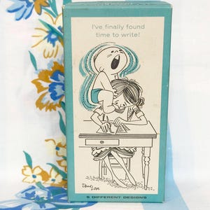 May include: A teal box with a cartoon illustration of a baby standing on a person's back while they write at a desk. The text on the box reads "I've finally found time to write!" and "5 DIFFERENT DESIGNS".
