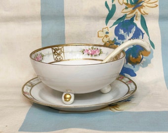 May include: A white porcelain bowl with gold trim and floral designs, sitting on a matching saucer with a small white ladle. The bowl has three feet and is decorated with pink roses and gold accents.
