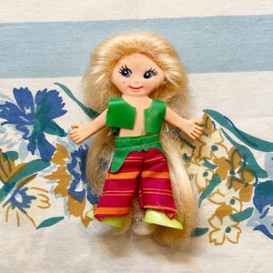 May include: A small plastic doll with long blonde hair, blue eyes, and a smiling face. The doll wears a green vest and red and orange striped pants. The doll's shoes are light green.
