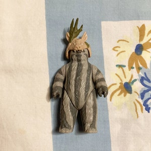 May include: A grey and brown furry action figure with a leafy headpiece. The figure has a striped pattern and is standing on a white and blue patterned surface.
