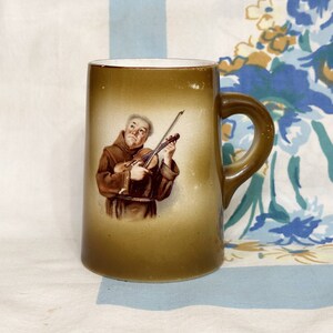 May include: A brown ceramic mug with a white interior features a vintage illustration of a man playing a violin. The man is wearing a brown robe and has a white beard.
