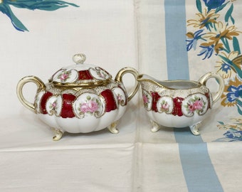 May include: A vintage porcelain sugar and creamer set with a red and gold floral design. The set features a sugar bowl with a lid and a creamer with a handle.