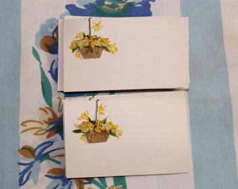 May include: Two blank white note cards with a yellow flower design. The flowers are in a basket and are hanging from a gold clip.