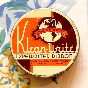 May include: A vintage round tin container for typewriter ribbon. The tin is orange and brown with the words "Klean-Write Typewriter Ribbon" and "Sold Round The World" printed on it. The tin has a globe graphic and the company name "Frankel Carbon & Ribbon Company, Denver, Detroit".