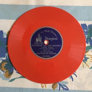 May include: A bright orange vinyl record with a blue label featuring Walt Disney's Disneyland logo and the text "Westward Ho, The Wagons! Theme Song, Part II, The Sandpipers, Mitch Miller and Orchestra, D308B".