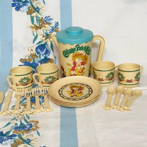 May include: A vintage Cabbage Patch Kids tea set in cream and teal. The set includes a pitcher, cups, plates, spoons, knives, and forks. The plates and pitcher feature the Cabbage Patch Kids logo and character illustrations.
